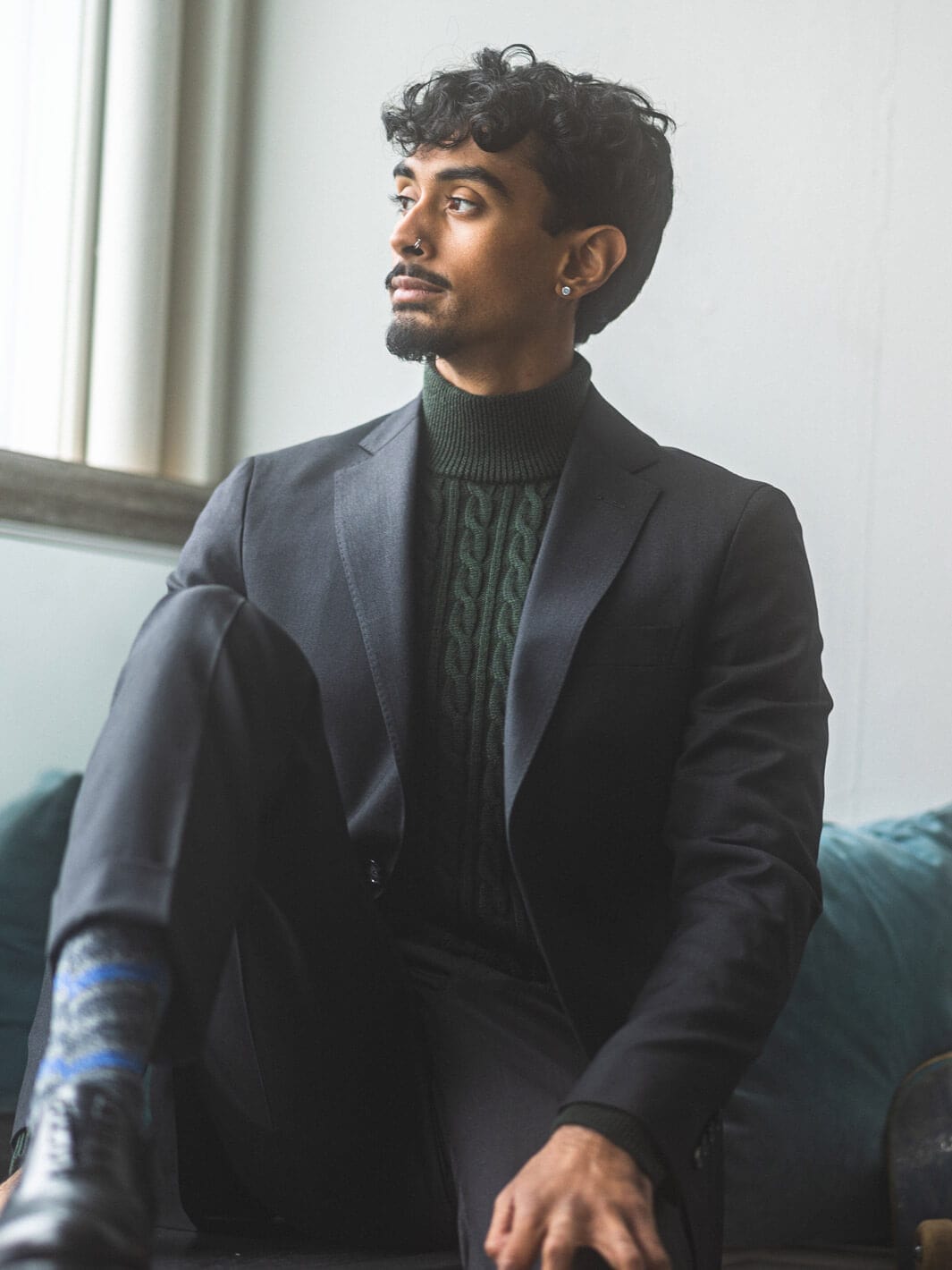 Man sitting on green couch with foot elevated on surface, looking out the window while wearing Anatoly's Drago Natural Stretch Suit in charcoal